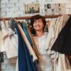 Joyful young ethnic lady smiling with closed eyes while standing amidst stack of clothes hanging on rack in fashion boutique