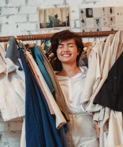 Joyful young ethnic lady smiling with closed eyes while standing amidst stack of clothes hanging on rack in fashion boutique