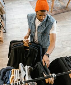 Man examines clothing in boutique wearing denim vest and orange beanie.