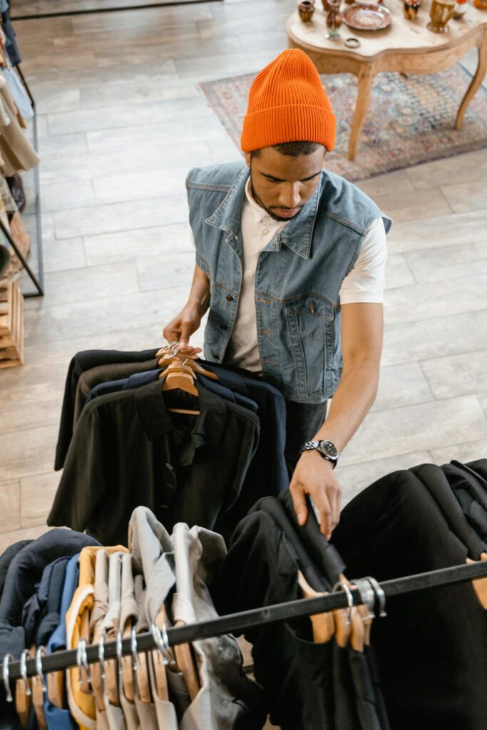 Man examines clothing in boutique wearing denim vest and orange beanie.